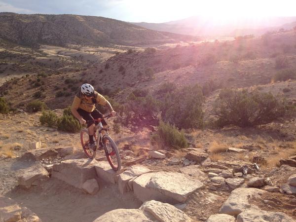 A mountain biker navigating rocky terrain on a trail with a scenic view of rolling hills and a sunset in the background. The cyclist is wearing a yellow shirt and helmet, skillfully maneuvering down a rocky slope. Kokopelli Area Trails mountain bike trail.