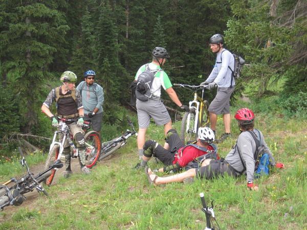 A group of five mountain bikers gathered on a grassy area surrounded by trees, some sitting on the ground and others standing with bicycles. They are dressed in casual cycling attire, wearing helmets and discussing their ride. A mix of bikes can be seen on the ground nearby. Trail 401 mountain bike trail.