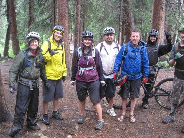 A group of six mountain bikers stands together in a forested area after a ride, wearing helmets and cycling gear. They are smiling and posing for the camera, with a mix of rain jackets and casual biking attire. A bicycle is parked beside one of the riders, and the ground is wet, indicating recent rain. The background features tall trees and a natural, wooded setting. Trail 401 mountain bike trail.