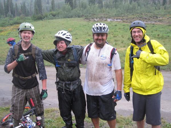 Four mountain bikers pose for a photo after an adventurous ride, smiling and showing signs of being muddy and wet. They are wearing helmets and various biking attire, standing together in a grassy area with trees in the background, exuding a sense of camaraderie and excitement. Trail 401 mountain bike trail.