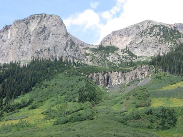 A scenic mountain landscape featuring rugged cliffs and peaks, lush green foliage in the foreground, and patches of colorful wildflowers. The sky is partly cloudy, with bright blue visible among the clouds. A small waterfall can be seen cascading down the mountainside. Trail 401 mountain bike trail.
