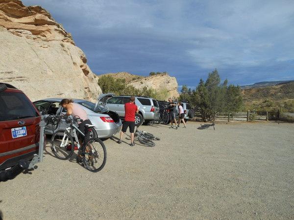 A group of people preparing for a biking trip at a gravel parking area near a rocky landscape. Several cars are parked, with some individuals unloading bicycles and gear from the trunks. A few trees and a wooden fence are visible in the background under a partly cloudy sky. Rustlers Loop mountain bike trail.