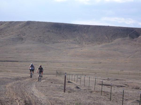 Two cyclists ride on a dirt path through a vast, arid landscape, with a hilly terrain in the background. The scene is characterized by dry, barren earth and sparse vegetation under a cloudy sky. A fence lines the path, emphasizing the open space around them. Gunnison River Bluffs Trail System mountain bike trail.