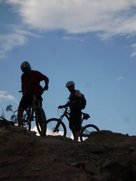Two mountain bikers are silhouetted against a blue sky as they navigate a rocky terrain. One biker is adjusting their bike while the other stands nearby, both wearing helmets and focused on the path ahead. Lunch Loops mountain bike trail.