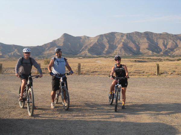 Three mountain bikers posing on a gravel path with a scenic backdrop of rugged mountains and open terrain. They are wearing helmets and biking gear, with two men and one woman smiling at the camera. The sun is shining, indicating a clear day. Joe's Ridge mountain bike trail.