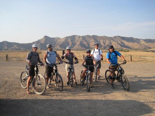 A group of six mountain bikers stands together in a sandy area, wearing helmets and casual cycling attire. They pose with their bikes against a backdrop of rugged mountains under a clear sky. Joe's Ridge mountain bike trail.