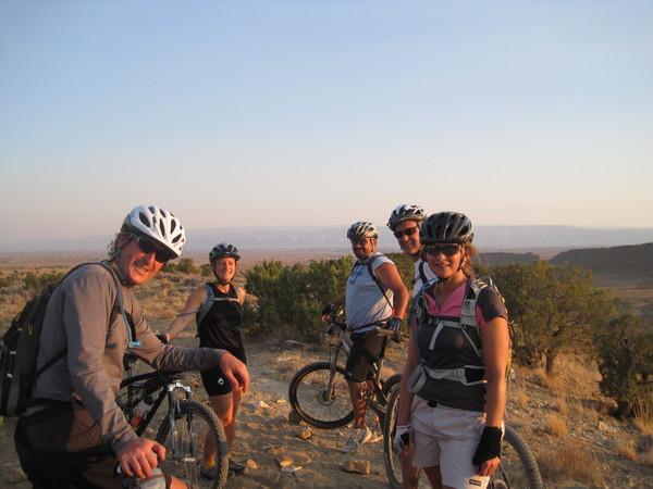 A group of five mountain bikers poses for a photo on a rocky outcrop with a scenic view in the background. They are wearing helmets and biking gear, smiling at the camera, with bicycles nearby. The landscape features dry terrain and sparse vegetation under a clear sky during sunset. Joe's Ridge mountain bike trail.