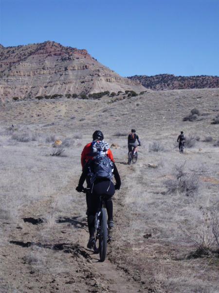 A group of mountain bikers riding along a dirt trail in a hilly desert landscape. The first biker, seen from behind, is wearing a black helmet and a bright red top, while others are visible further down the trail with a backdrop of rocky hills under a clear blue sky. Sparse vegetation and dry grass cover the ground. 18 Road Trails / North Fruita Desert mountain bike trail.