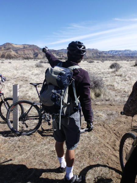 A person in a cycling helmet and outdoor gear is standing with their back to the camera, pointing towards a distant mountain range in a dry, open landscape. They are wearing a large backpack and shorts, with bicycles parked nearby. The sky is clear and blue, suggesting a sunny day. 18 Road Trails / North Fruita Desert mountain bike trail.