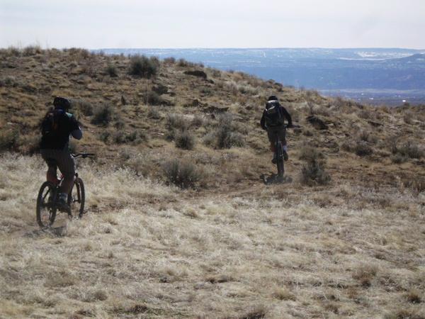 Two cyclists riding mountain bikes on a grassy hillside, with mountains and a blue sky visible in the background. The terrain appears rugged, and both riders wear helmets. 18 Road Trails / North Fruita Desert mountain bike trail.
