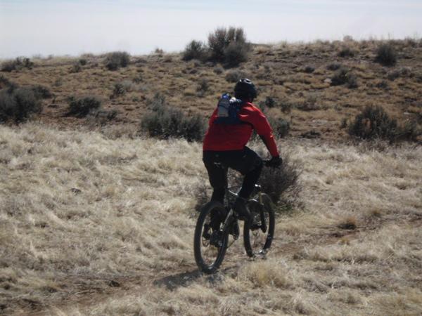 A person riding a mountain bike through a grassy landscape, wearing a red jacket and a black helmet. They are heading away from the camera, navigating a trail among sparse vegetation and hills. 18 Road Trails / North Fruita Desert mountain bike trail.