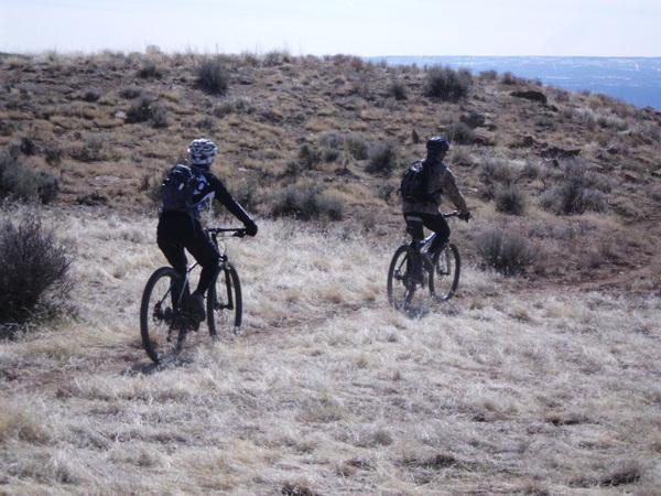 Two mountain bikers ride along a dirt path through a grassy landscape with sparse vegetation. The terrain is slightly hilly, and the sky is clear, indicating a bright day. The bikers are wearing helmets and cycling gear, with one cyclist in a blue and black outfit and the other in muted colors. 18 Road Trails / North Fruita Desert mountain bike trail.