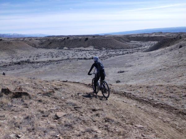 A person riding a mountain bike down a steep, rocky trail in a dry, open landscape with rolling hills and sparse vegetation under a clear blue sky. 18 Road Trails / North Fruita Desert mountain bike trail.