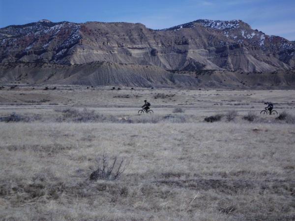 Two mountain bikers ride through a vast, dry landscape with rocky hills in the background. The terrain is mostly beige and brown, with sparse vegetation. Snow caps the peaks of the hills, and the sky is clear with soft clouds. 18 Road Trails / North Fruita Desert mountain bike trail.