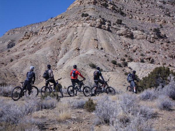 A group of five mountain bikers stands on a trail, overlooking a rugged landscape with steep, rocky hills in the background. The terrain is dry and sparse, with some low shrubs. The bikers are wearing helmets and backpacks, ready for an outdoor adventure. 18 Road Trails / North Fruita Desert mountain bike trail.