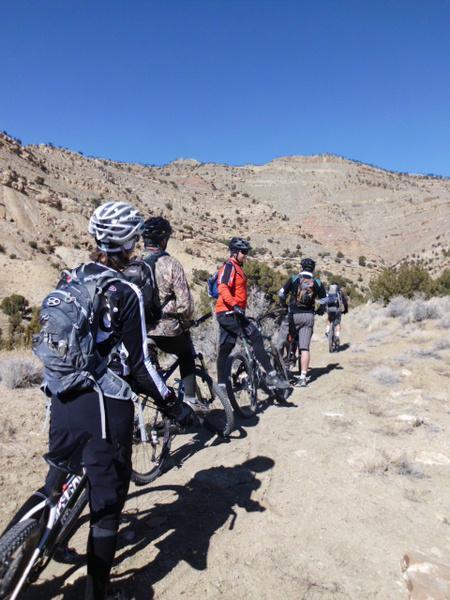 A group of mountain bikers pause along a trail in a rugged, arid landscape. The riders, wearing helmets and backpacks, are positioned in a line, facing away from the camera. The background features rocky hills and clear blue skies, creating a scenic outdoor adventure atmosphere. 18 Road Trails / North Fruita Desert mountain bike trail.