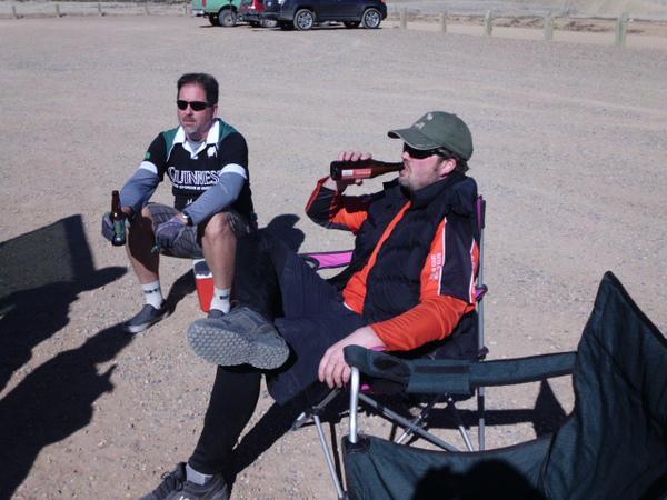 Two men sitting outdoors on a gravel surface. One man in the foreground is wearing a black vest and an orange long-sleeve shirt, holding a drink, and relaxed in a camping chair. The other man, in the background, is dressed in a green and black shirt with sunglasses, sitting in a similar chair with a beer bottle in his hand. A parked car is visible in the background. 18 Road Trails / North Fruita Desert mountain bike trail.