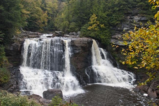 A scenic waterfall cascading over rocky formations into a calm pool, surrounded by lush greenery and autumn-colored trees. Blackwater Falls State Park mountain bike trail.