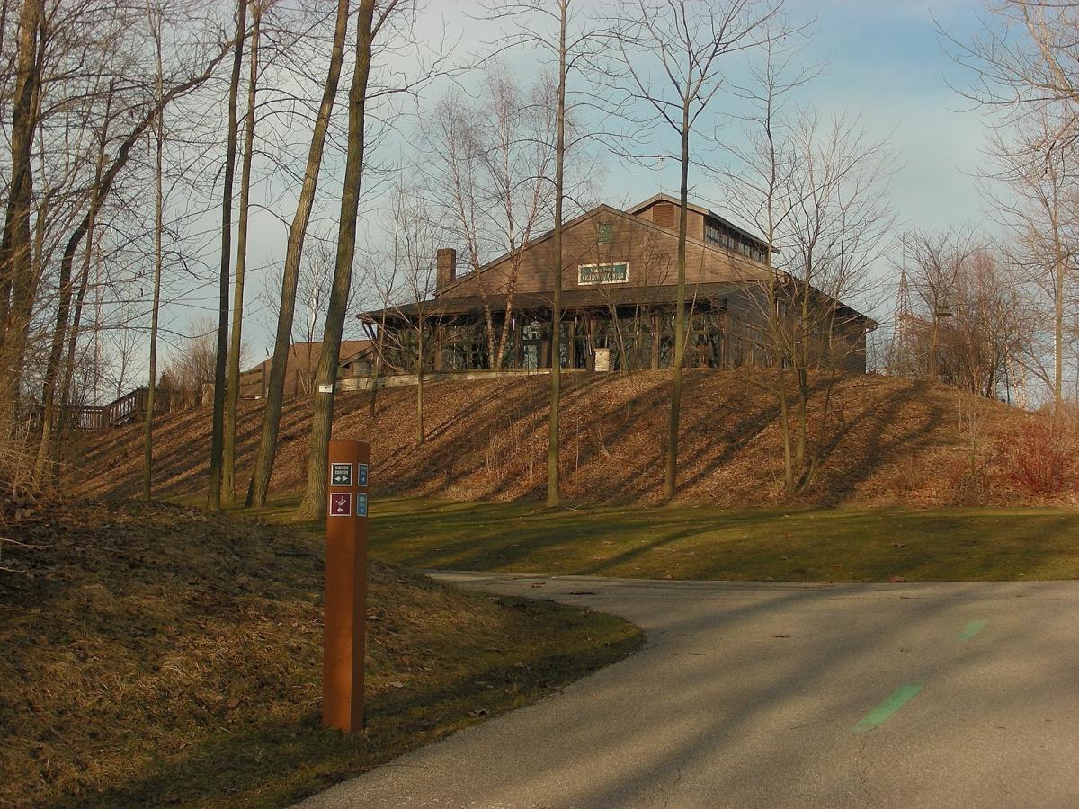 A view of a large, wooden building situated on a hill, surrounded by bare trees and grassy areas. A pathway leads up to the structure, with a signpost nearby indicating marked trails and visitor information. The sky is partly cloudy, and the scene is set in a natural environment. Ohio And Erie Canal mountain bike trail.