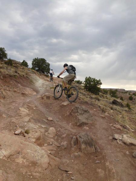 A person riding a mountain bike down a rocky trail, with a cloudy sky in the background. Another cyclist is visible in the distance on the same path, surrounded by sparse vegetation. Lunch Loops mountain bike trail.