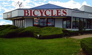 Exterior view of a bicycle shop featuring large windows, a blue roof, and the word "BICYCLES" prominently displayed in red. There is a bicycle mounted on the roof and well-maintained green shrubs in the foreground, with a clear blue sky visible in the background.