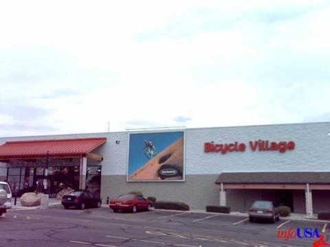 Exterior view of Bicycle Village, a bicycle shop with a prominent red awning and large signage. A vibrant advertisement featuring a cyclist on a mountain trail is displayed on the building