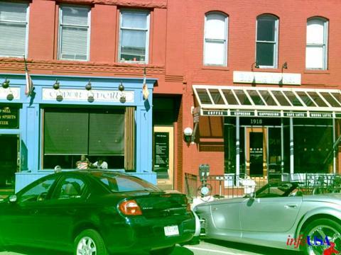 A bustling street scene featuring two adjacent buildings with red brick facades. The left building has a black awning and large windows, possibly a restaurant or café. The right building, also with large windows, displays the year "1918" prominently. Two parked cars, one black and one silver, are visible in front of the buildings. The image has a greenish tint.
