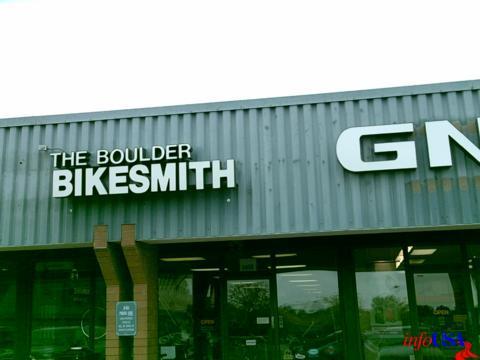 Signage for "The Boulder Bikesmith" displayed on the exterior of a building, with a gray corrugated metal facade. The sign features bold, black lettering, and there is a second sign for "GN" nearby. Part of the building's entrance is visible.