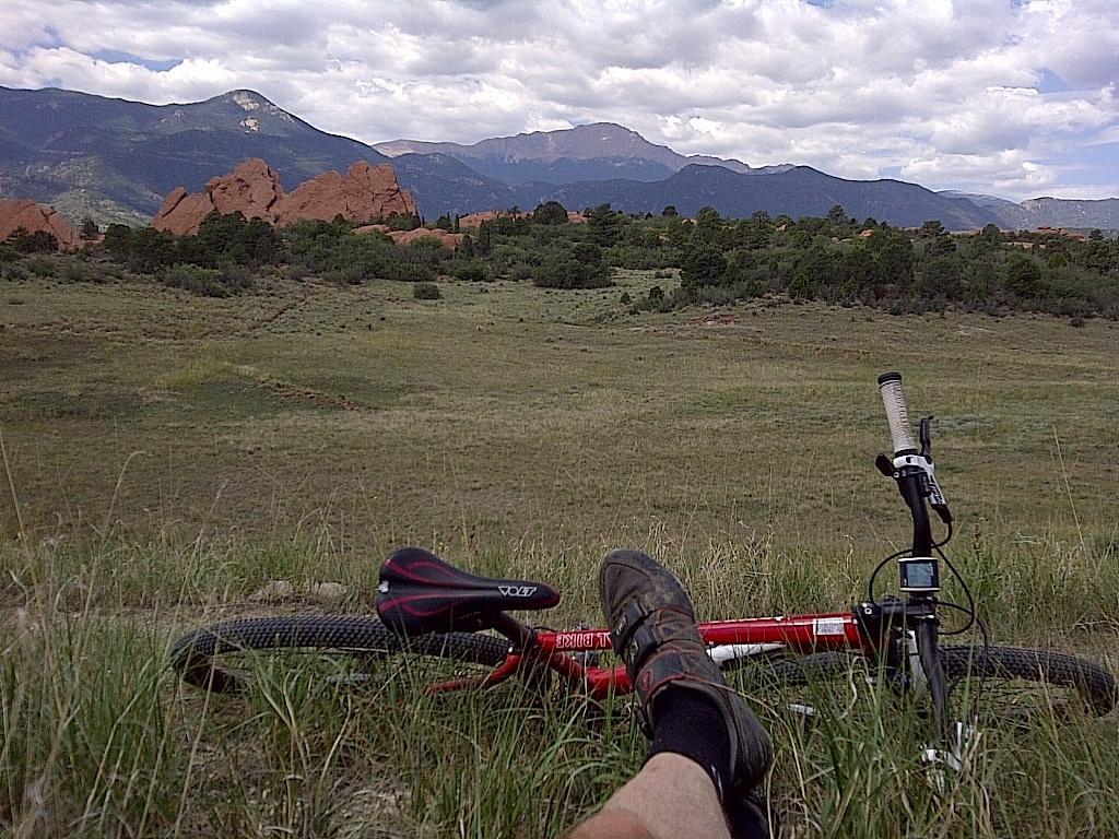 A mountain biker's feet are resting on the grass, with a red mountain bike beside them, against a backdrop of rocky formations and distant mountains under a partly cloudy sky. Garden of the Gods: Ute Trail mountain bike trail.