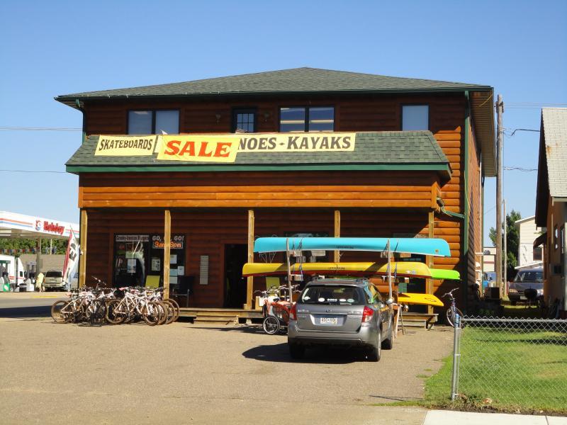 A wooden building with a green roof, featuring a large sign that reads "SALE" along with the words "SKATEBOARDS" and "CANOE - KAYAKS." In front of the store, there are colorful kayaks displayed on a rack, along with several bicycles parked nearby. A car is parked in the driveway, and the background includes a gas station and neighboring buildings. The scene is set on a sunny day with clear blue skies.