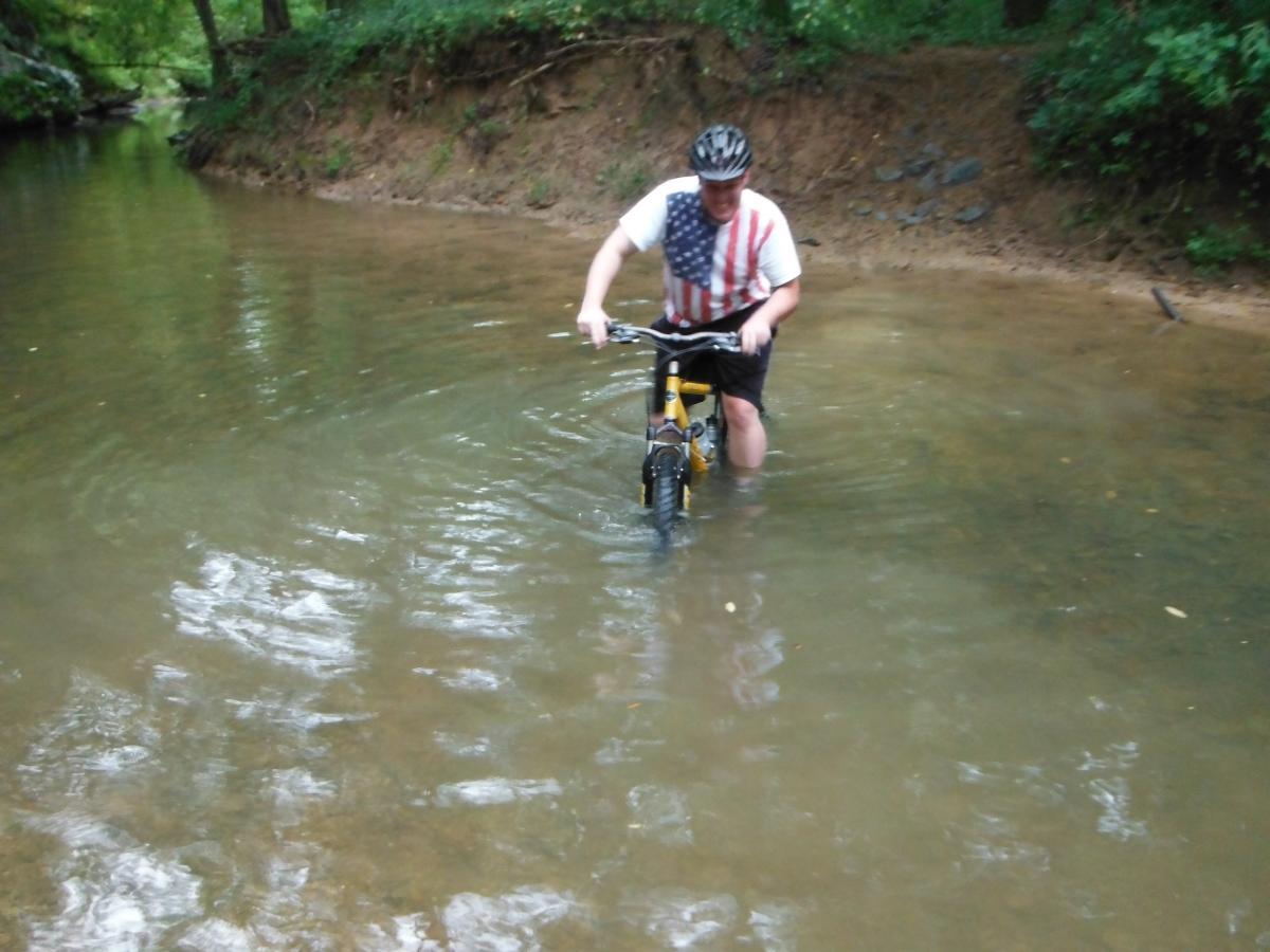 A person wearing a helmet and an American flag T-shirt is walking a bicycle through shallow water in a wooded area. The water is clear and reflects the surrounding greenery, while the bank is lined with dirt and small rocks. Dry Creek mountain bike trail.