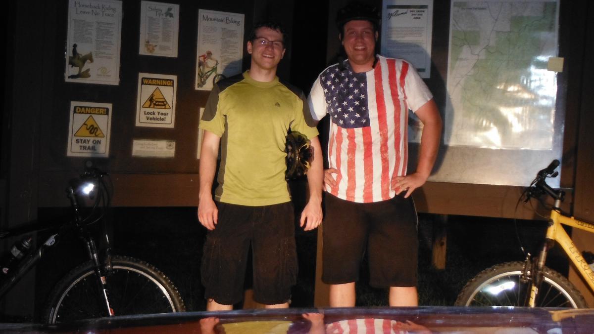 Two men stand smiling in front of a trail signage board, showcasing information about mountain biking. One man wears a green shirt and holds a helmet, while the other sports a white shirt with an American flag design. Their bicycles are parked nearby, and the setting appears to be in a natural outdoor area, with some trees and grass visible. Dry Creek mountain bike trail.