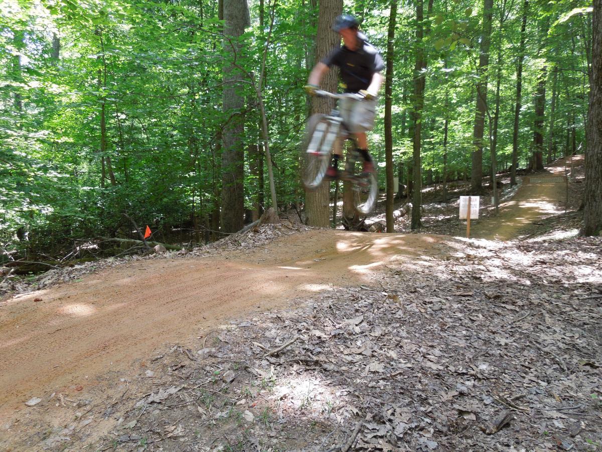 A mountain biker catching air off a dirt jump on a wooded trail, surrounded by green trees and underbrush, with a partially visible trail in the background. Fountainhead Regional Park mountain bike trail.