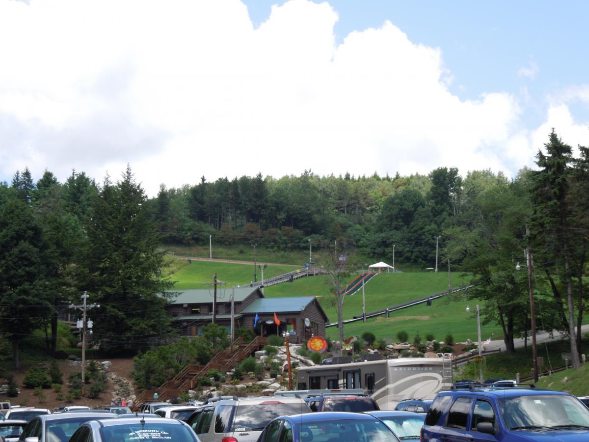 A parking lot filled with vehicles, with a recreational facility in the background. The facility features a green hill for outdoor activities, including a sloped track and colorful slides. Surrounding trees provide a natural backdrop under a partly cloudy sky. Seven Springs mountain bike trail.