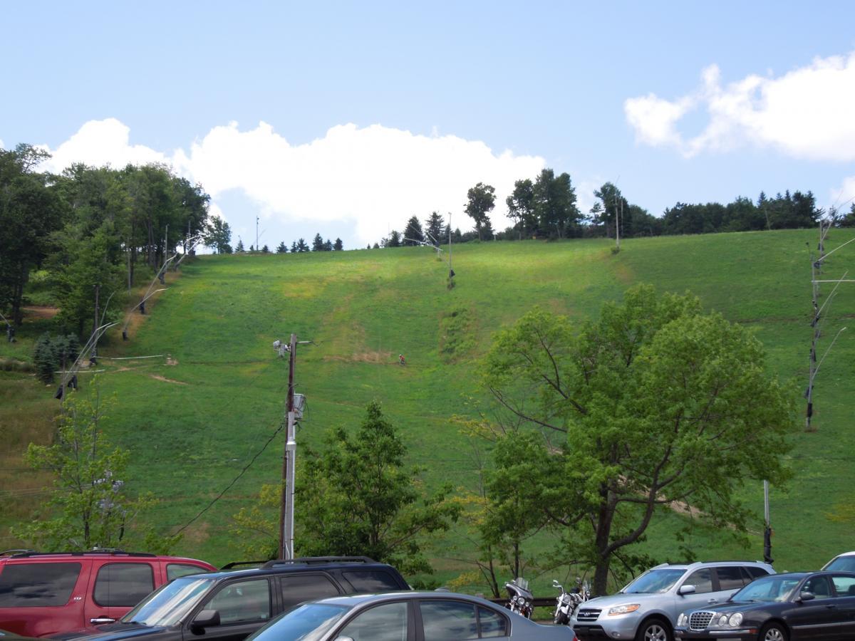 A scenic view of a grassy slope with a ski lift and trees in the background, taken on a clear day with a blue sky and fluffy clouds. In the foreground, parked cars are visible along with some greenery. Seven Springs mountain bike trail.