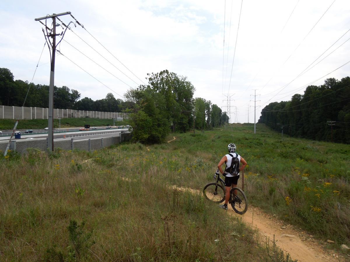 A cyclist stands on a dirt path, overlooking a grassy area with wildflowers. In the background, a highway runs parallel, flanked by a sound barrier and power lines stretching into the distance. Trees line the area, creating a mix of urban and natural scenery under a cloudy sky. Wakefield mountain bike trail.