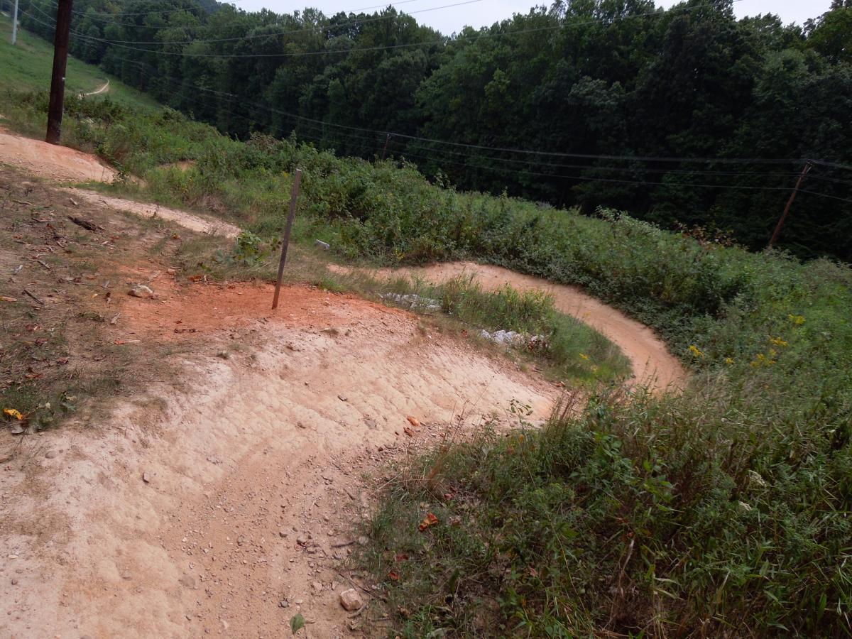 A winding dirt path cuts through a green hillside, bordered by tall grass and shrubs. Electrical poles run along the left side, with trees visible in the background. The path is marked by a sandy texture with areas of reddish soil, suggesting recent use for biking or hiking. Wakefield mountain bike trail.
