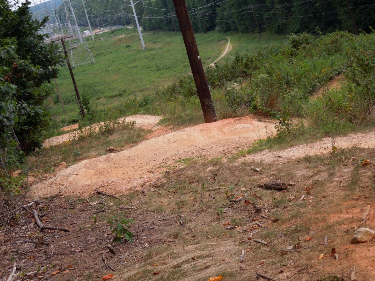 A dirt trail winding down a hillside surrounded by greenery, with power lines in the background. The path shows signs of erosion, with scattered rocks and debris along the edges. Wakefield mountain bike trail.