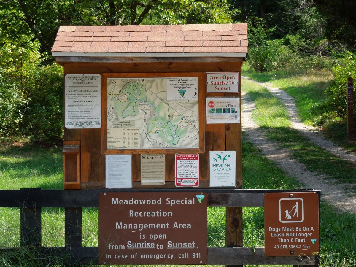 A wooden information kiosk in a natural setting, displaying a map and various notices about the Meadowood Special Recreation Management Area. The signage indicates that the area is open from sunrise to sunset, provides guidelines for visitors including dog leash regulations, and includes information about invasive plants and bird habitats. A narrow dirt path can be seen leading into the wooded area. Meadowood mountain bike trail.