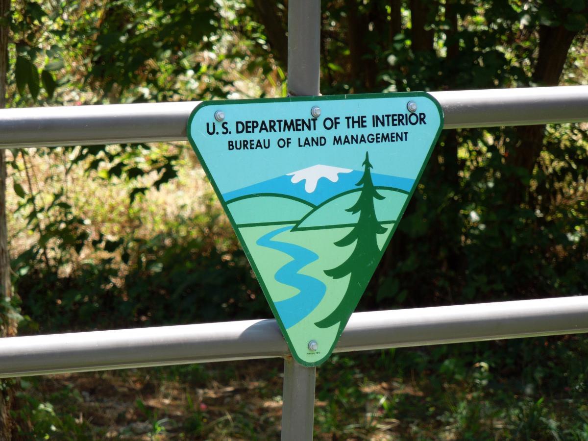 A triangular sign mounted on a metal fence, featuring the text "U.S. DEPARTMENT OF THE INTERIOR BUREAU OF LAND MANAGEMENT" along with an illustration of a river, mountains, and evergreen trees against a blue sky. Surrounding foliage is visible in the background. Meadowood mountain bike trail.