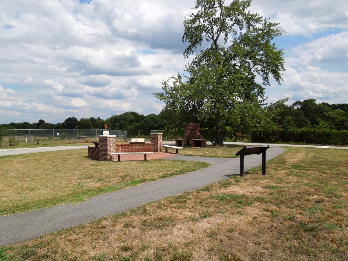A grassy area with a brick seating structure, a large tree, and signs along a walking path. The background features a fenced area with trees and clouds in the sky. Laurel Hill Park mountain bike trail.
