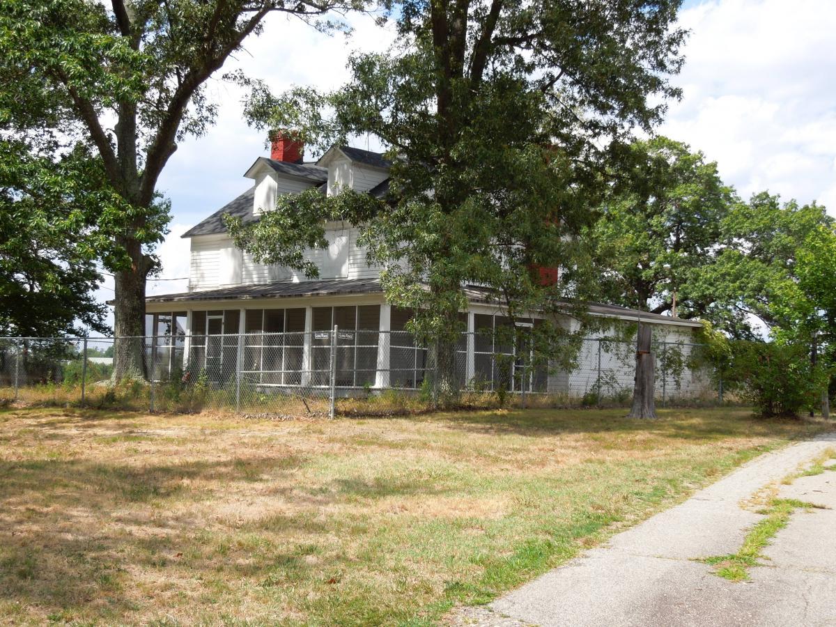 An old two-story house surrounded by trees and a chain-link fence, featuring a porch with a screened-in area. The yard is grassy and dry, with a gravel path leading towards the house, under a partly cloudy sky. Laurel Hill Park mountain bike trail.