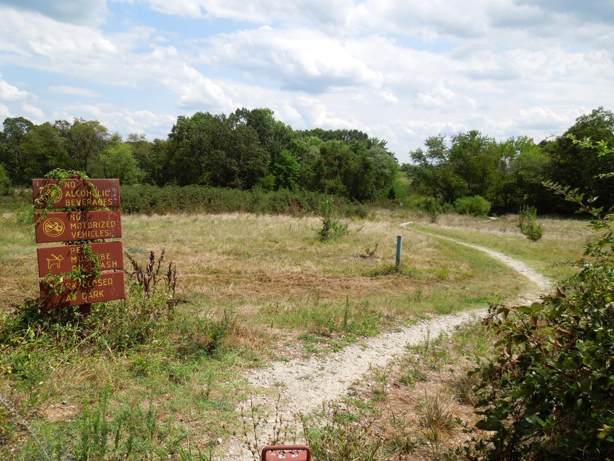 A pathway winding through an open field, bordered by trees in the background. On the left, a sign wooden sign displays rules including prohibitions on alcoholic beverages and motorized vehicles, alongside pet regulations. The ground is dry and grassy, with sparse greenery. The sky is partly cloudy, suggesting a warm day. Laurel Hill Park mountain bike trail.