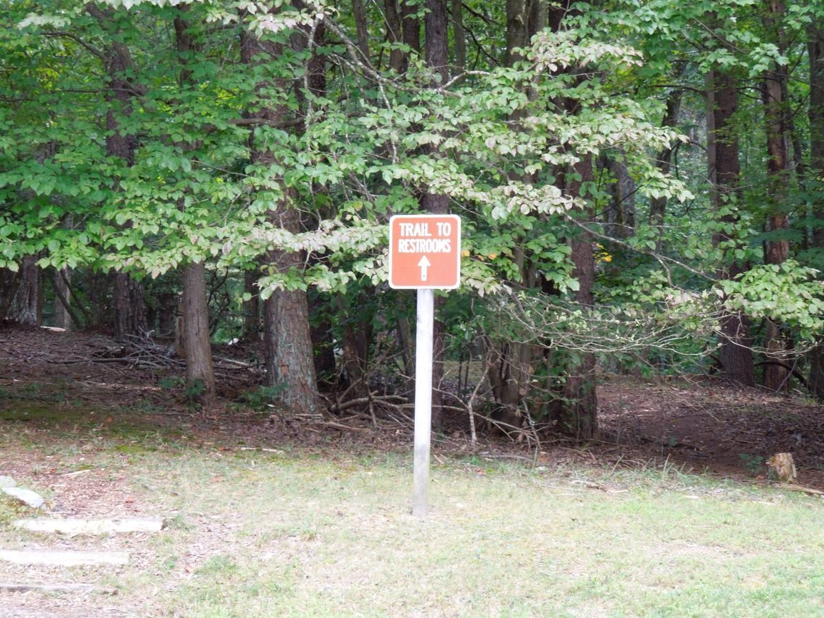 A sign indicating the trail to restrooms, positioned in a wooded area with dense green foliage. The sign features an arrow pointing in the direction of the restrooms. Fountainhead Regional Park mountain bike trail.