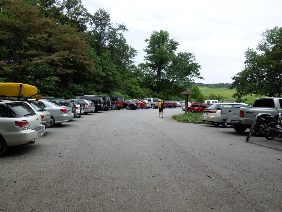 A parking lot filled with various cars, including several SUVs and sedans, with a kayaker's yellow kayak visible on one vehicle's roof. In the background, trees and a green landscape are visible under a cloudy sky. A person is walking along the edge of the parking area. Schaeffer Farms mountain bike trail.