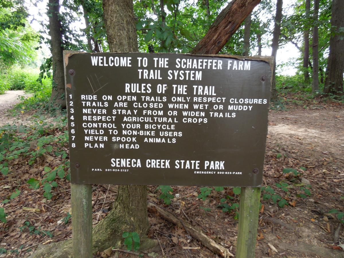 A wooden sign at the entrance of the Schaeffer Farm Trail System in Seneca Creek State Park, detailing the rules for trail use, surrounded by trees and a natural pathway. The rules include riding only on open trails, respecting closure signs, and being mindful of agricultural crops and other trail users. Schaeffer Farms mountain bike trail.
