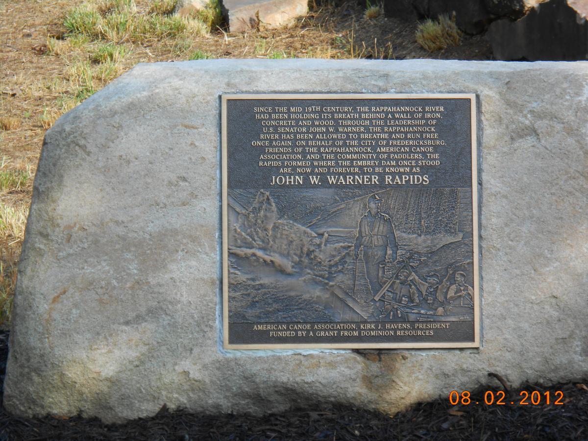 A bronze plaque mounted on a large stone commemorating the restoration of the Rappahannock River and renaming of the rapids to "John W. Warner Rapids." The plaque includes historical text discussing the significance of the river since the mid-19th century and acknowledges contributions from various organizations and individuals. The background features natural surroundings with grass and trees. The date "08.02.2012" is visible in the bottom corner. Rappahanock River Trail mountain bike trail.