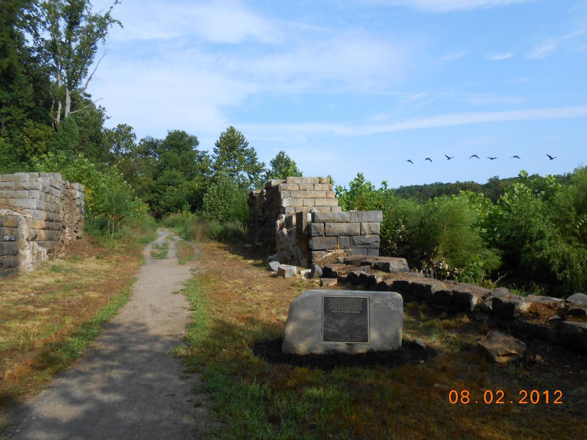 A dirt path leads through overgrown greenery, flanked by the remnants of stone structures on either side. In the foreground, a stone monument with an engraved plaque is visible, while a line of birds flies across a clear blue sky above the trees in the background. Rappahanock River Trail mountain bike trail.