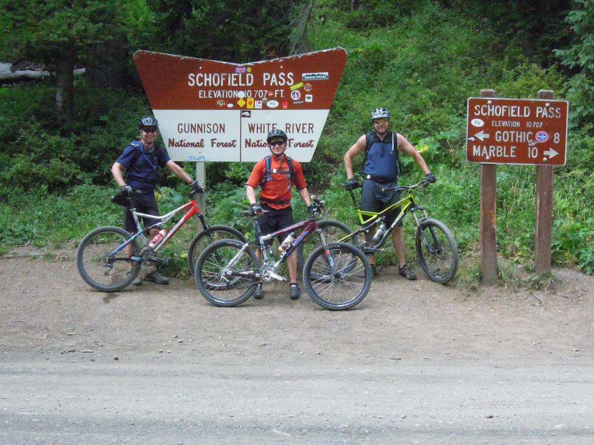 Three mountain bikers pose with their bikes at Schofield Pass, surrounded by lush greenery. A large sign in the background indicates the elevation of 10,707 feet and provides information about nearby trails in Gunnison and White River National Forests. Trail 401 mountain bike trail.