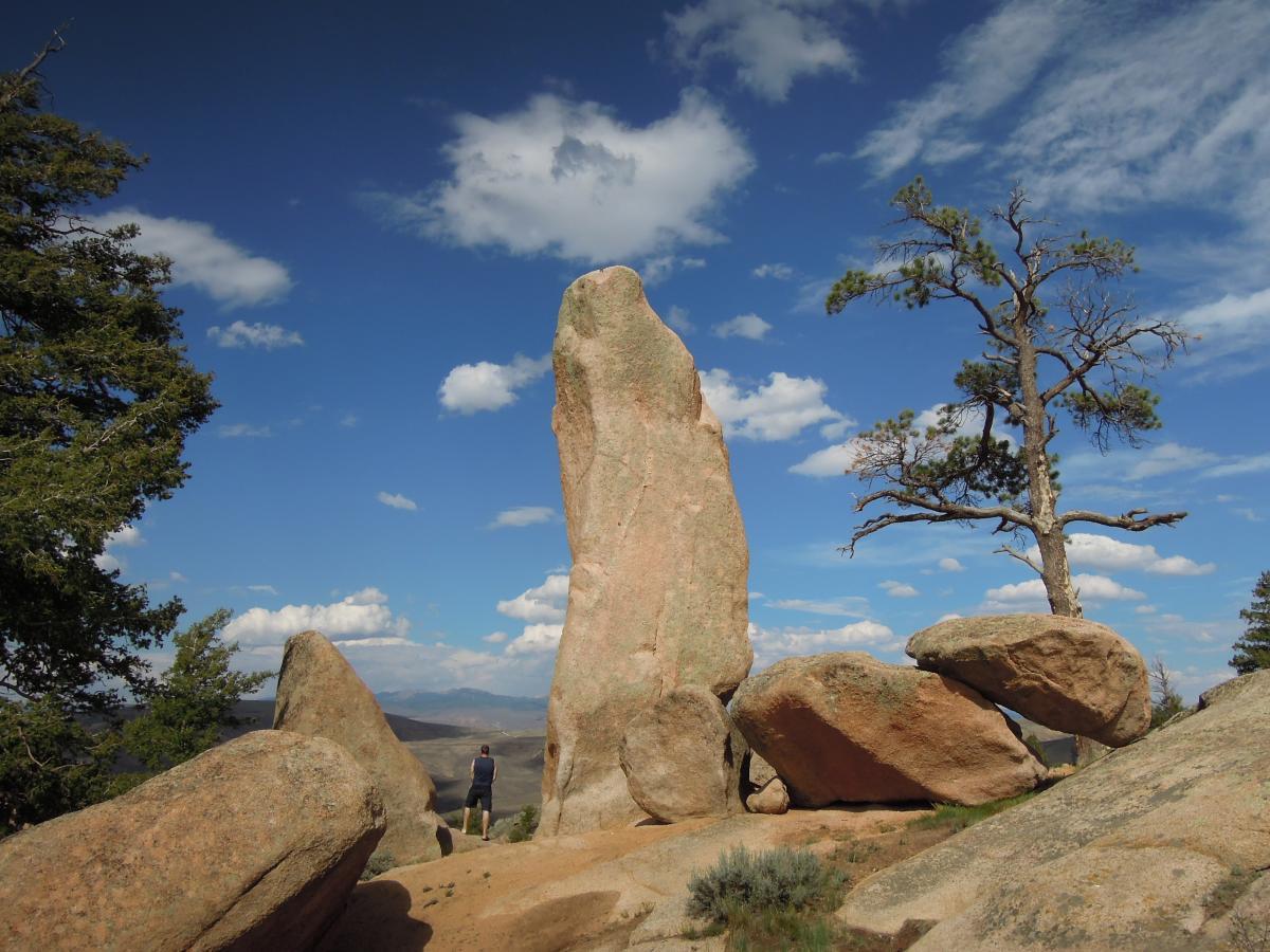 A person standing in front of a tall rock formation amidst boulders, with a tree on the right and a clear blue sky dotted with clouds above. The landscape is rocky, with rolling hills visible in the background. Hartman Rocks mountain bike trail.